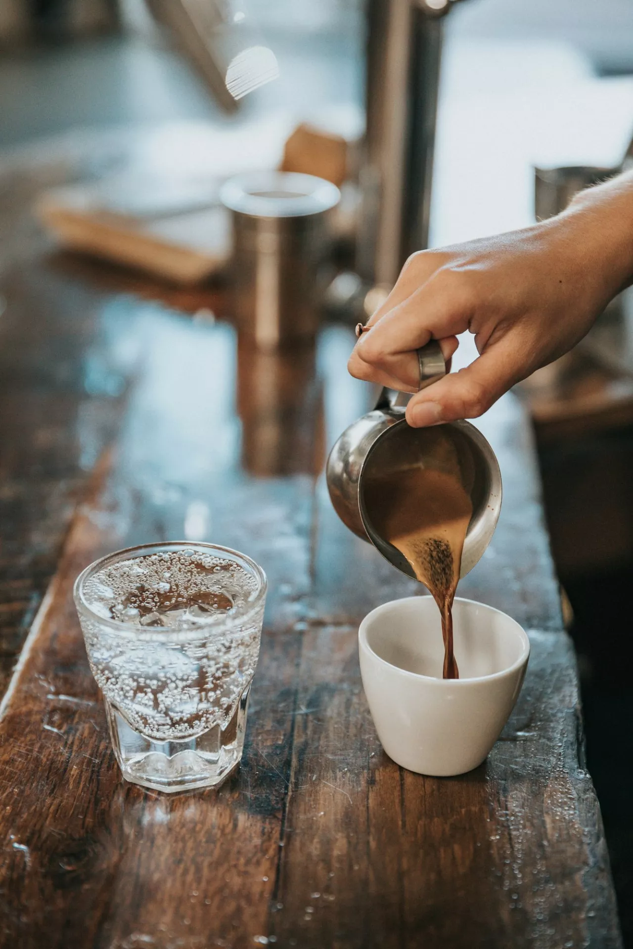 A hand is pouring coffee from a metal pitcher into a white cup on a wooden table, with a glass of sparkling water nearby.
