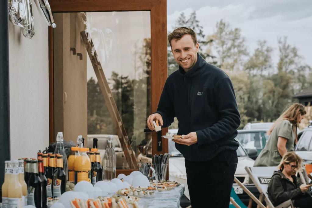 A man smiling while picking food from a table filled with drinks and snacks during an outdoor gathering.