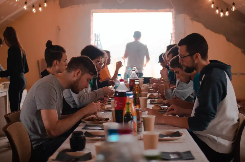 A group of people is seated around a long table, eating and socializing in a cozy, warmly lit room.