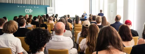 A large audience is attending a conference, listening to a speaker presenting at the front of the room.