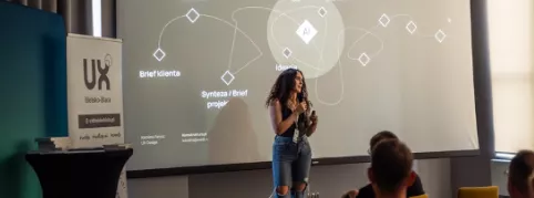 A speaker presents to an audience in front of a screen displaying a diagram.