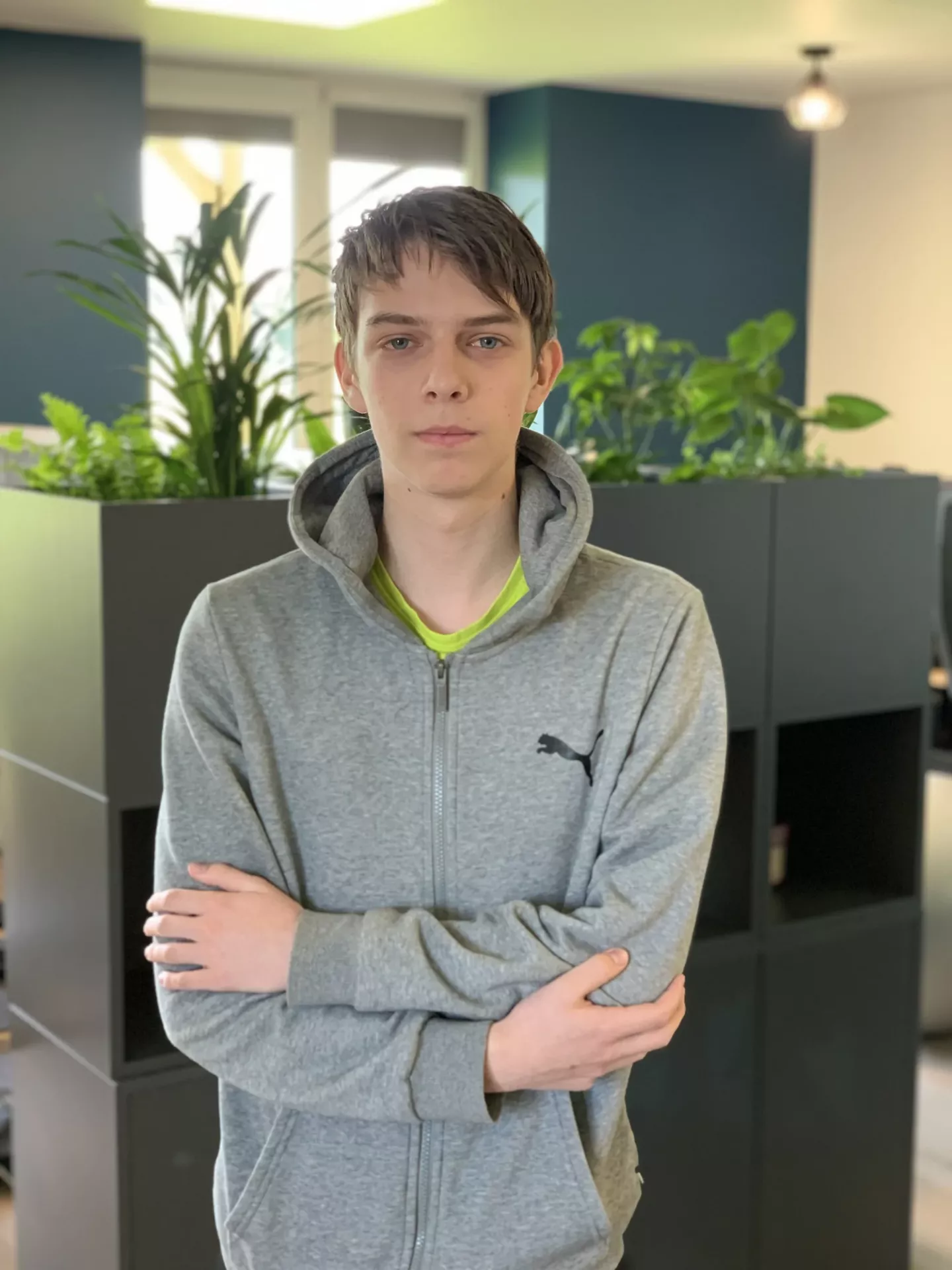 A person with short hair, wearing a gray hoodie, stands with arms crossed in an office with plants in the background.