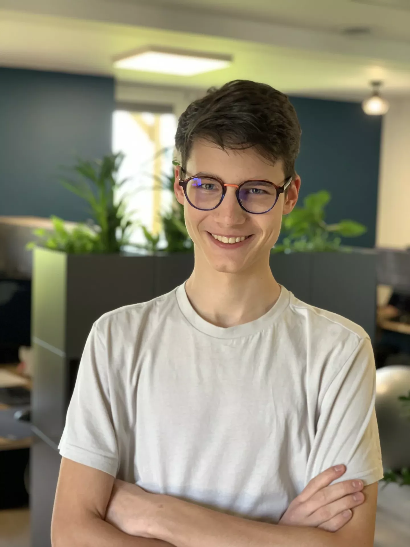 A handsome person with short hair and glasses, wearing a light T-shirt, stands with arms crossed in an office with plants.