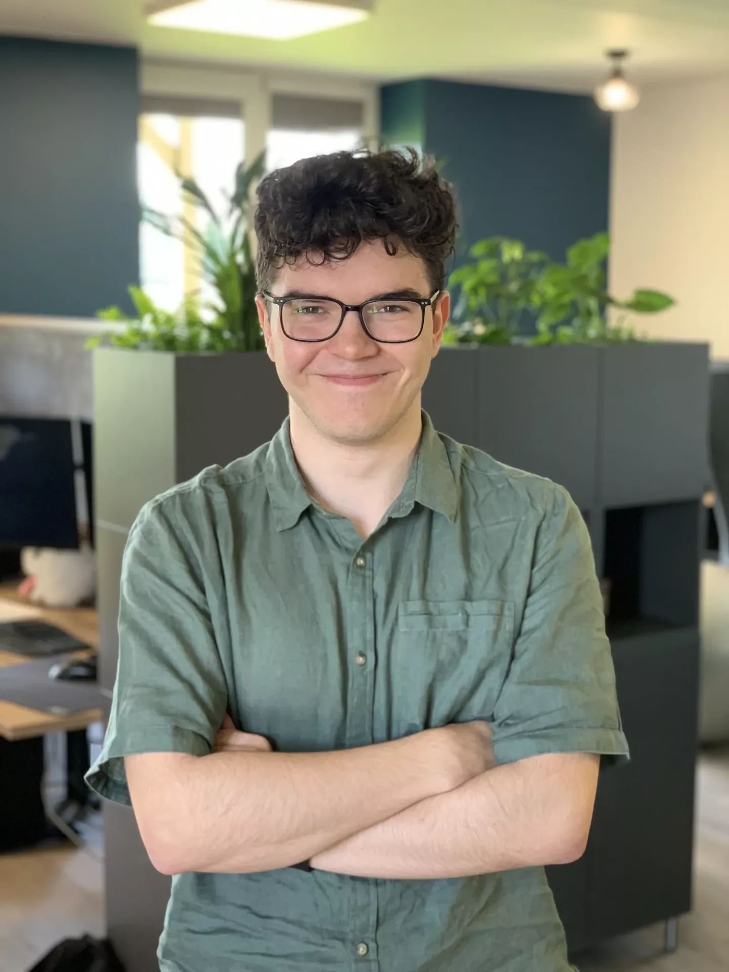 A person with curly hair and glasses, wearing a green shirt, stands with arms crossed in an office with plants.