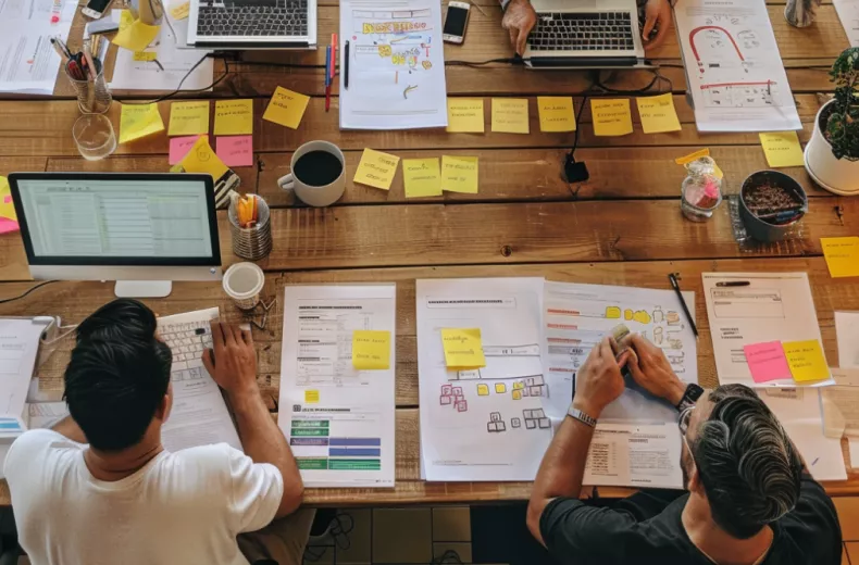 People are working at a table with laptops, papers, and sticky notes, collaborating on a project.
